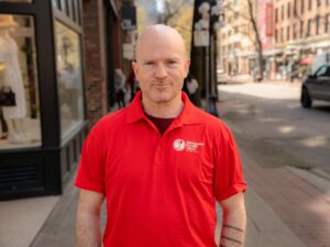 A man with a shaved head and a slight smile looks into the camera. He's wearing a red Vancouver Foodie Tours shirt