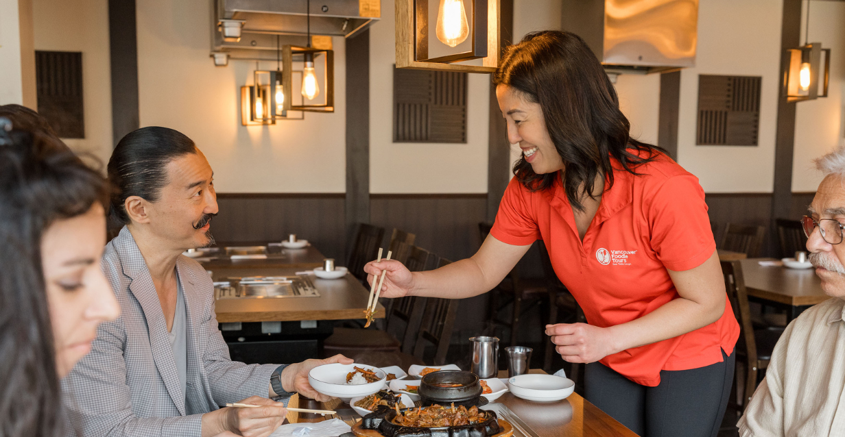 A foodie tour guide serves a dish with chopsticks to a guest who is smiling, with other guests gathered around a table
