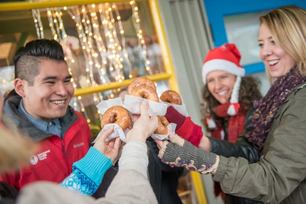 A group of tourists on a Vanoucver Festive Favourites Market Tour holding doughnuts in paper napkins and raising them for a cheer together with their local guide