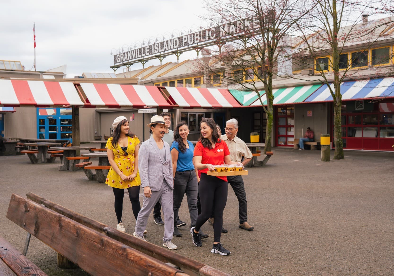 A group of people follow their guide with Granville Island PUblic Market in the background. They're all smiling as guide Manuela carries a tray of drinks.