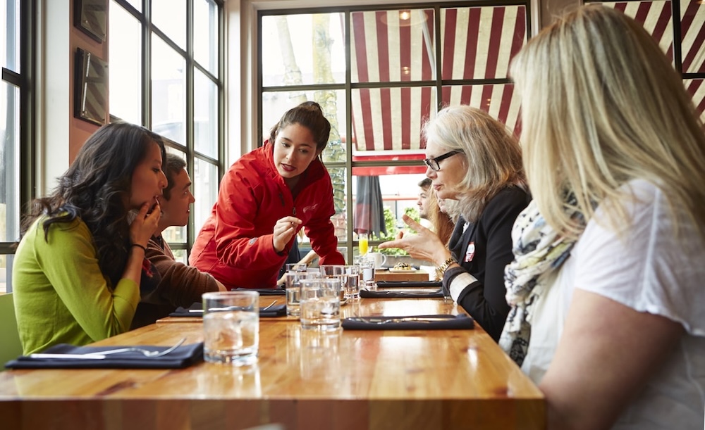 a tour guide standing at a table with her guests seated around, making sure that each one of them is taken care of and catered to