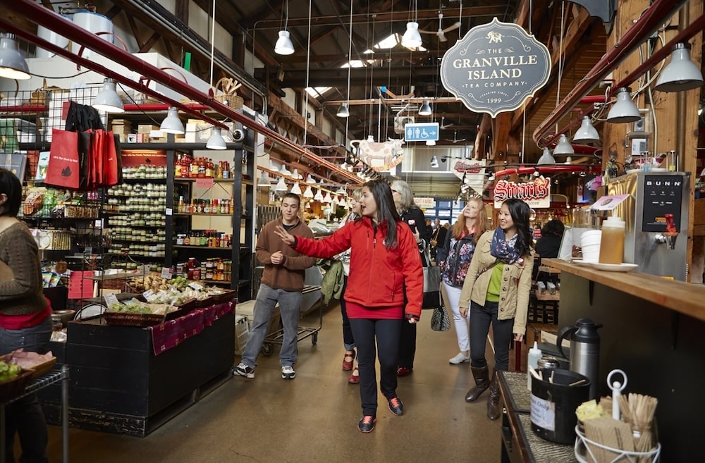 A Vancouver Foodie Tours guide in a red jacket leads a tour group through the Granville Island market