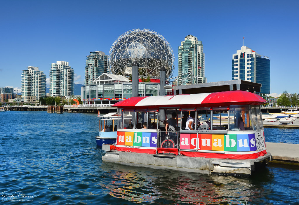 A small, colorful AquaBus ferry crossing False Creek with the Granville Island Public Market in the background.