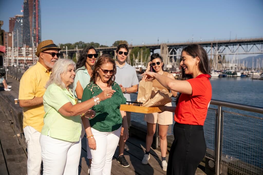 A Vancouver Foodie Tours guide in a red shirt hands out samples to a smiling group of tour participants on a sunny boardwalk with boats and a bridge in the background.