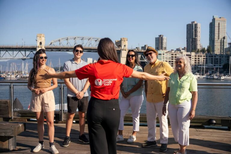 Vancouver Foodie Tours guide in a red logo t-shirt with arms outstretched, welcoming a small group of guests with Granville Island in the background.
