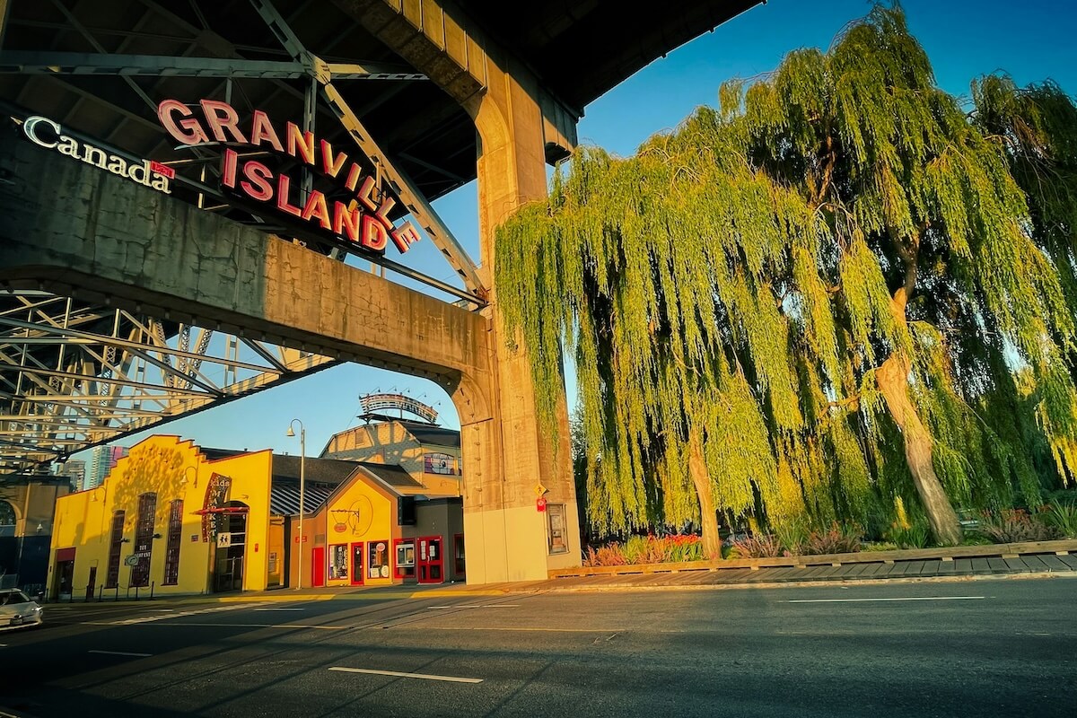 Granville Island entrance at evening with willow trees and colorful buildings under the bridge.