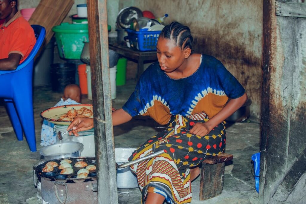 Woman cooking over a stove indoors, tending a pan of round fried breads, with kitchen items in the background.