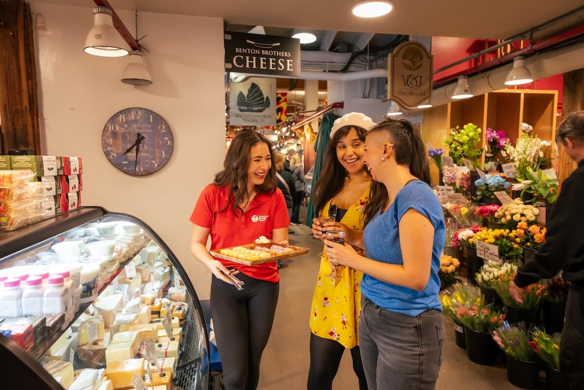 Vancouver Foodie Tours guide offering cheese samples to two guests at Benton Brothers Fine Cheese inside Granville Island Public Market.